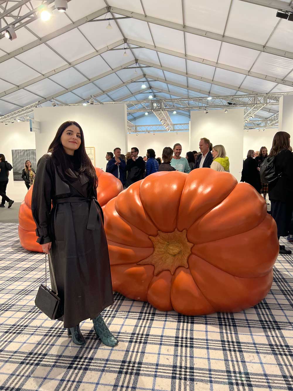 Amalia Nangeroni at an international art fair standing beside an oversized resin pumpkin sculpture — art advisory site visit