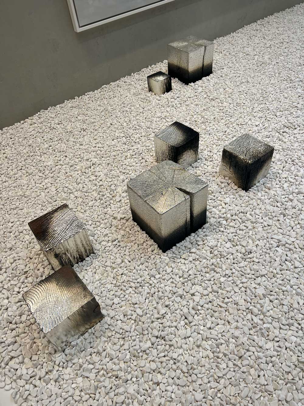Gallery floor installation of multiple burnished steel cube sculptures of varying sizes scattered on a white gravel bed — contemporary art advisory and collection