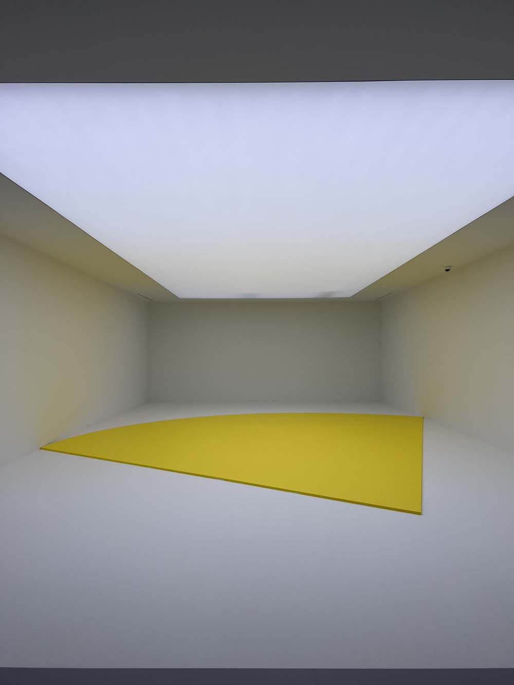 Minimalist contemporary art installation: white room with a large yellow wedge-shaped floor element under diffused zenithal light — art advisory and collection management