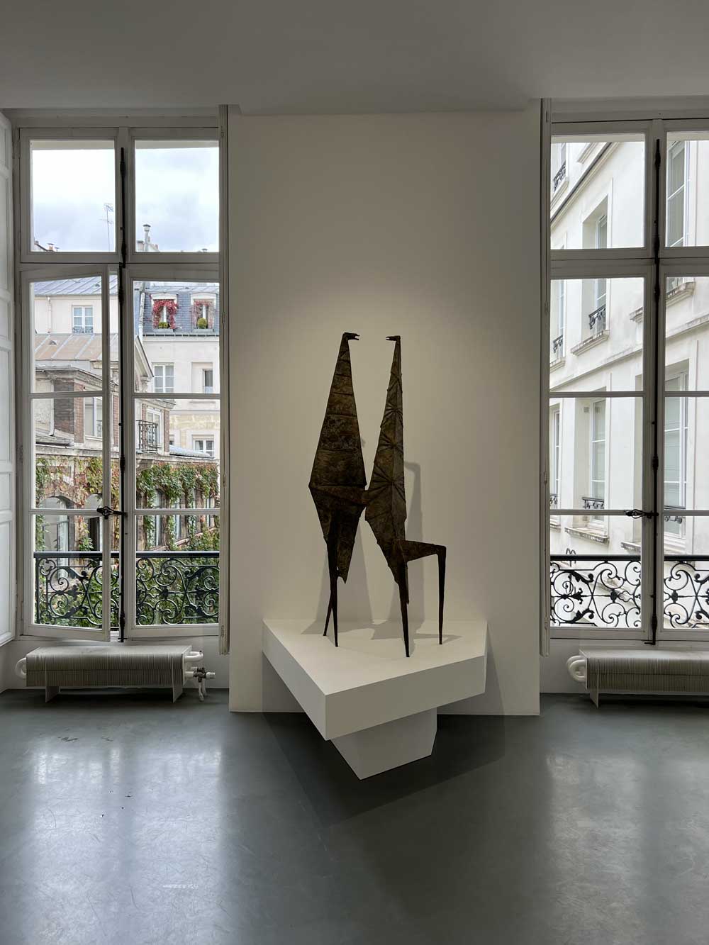 Two abstract bronze sculptures of elongated stylised figures on a plinth in a Paris gallery with Haussmann building views — contemporary art advisory