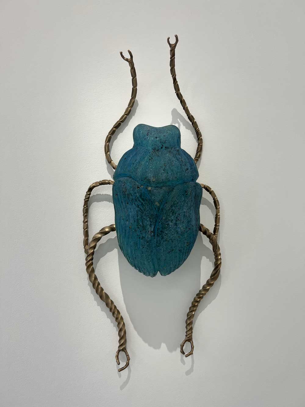 Sculptural wall work: oversized blue scarab beetle in ceramic with brass legs mounted on a white wall — contemporary art advisory