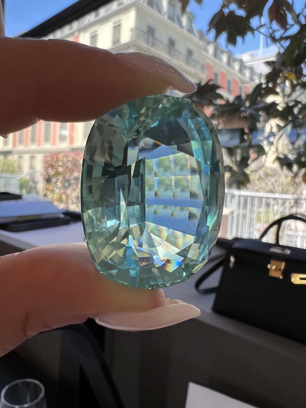 Large faceted light-green gemstone held up to the light with Geneva's historic waterfront visible in the background — international diamonds and jewelry advisory