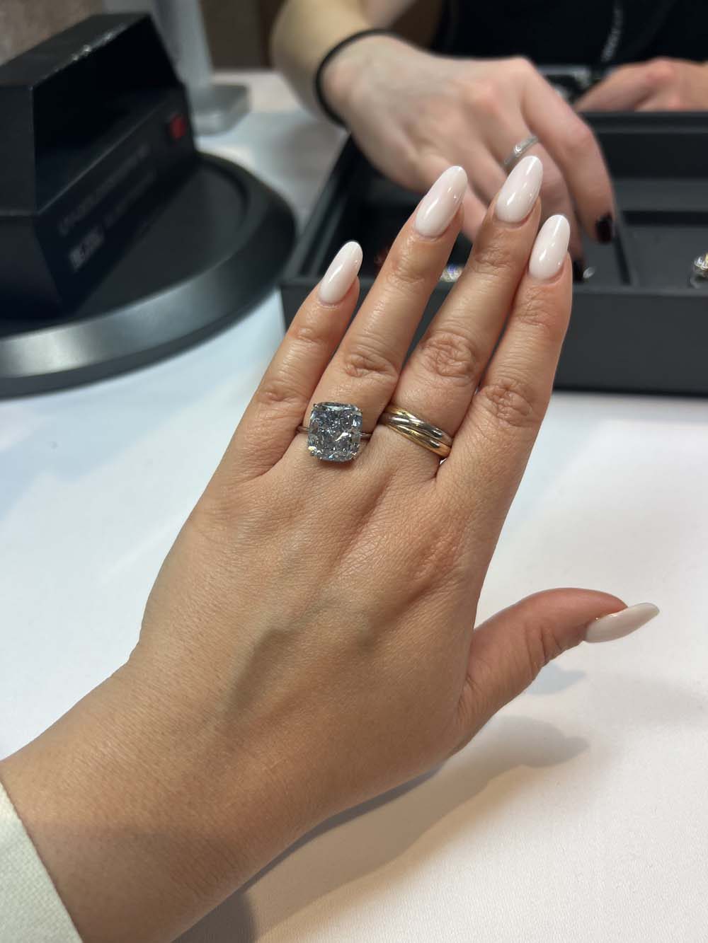 Large cushion-cut diamond solitaire ring tried on hand, with GIA grading equipment visible in the background — diamond advisory and valuation