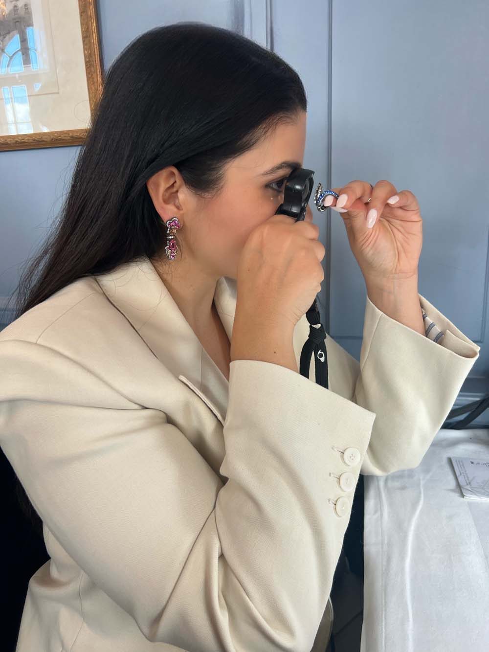 Amalia Nangeroni examining a gemstone with a jeweler's loupe, wearing ruby and diamond flower earrings — expert diamonds and jewelry advisory